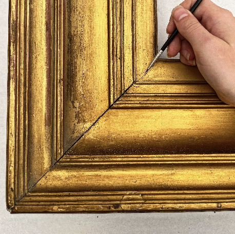 A restorer is using a paintbrush to retouch the gold on a gilded frame